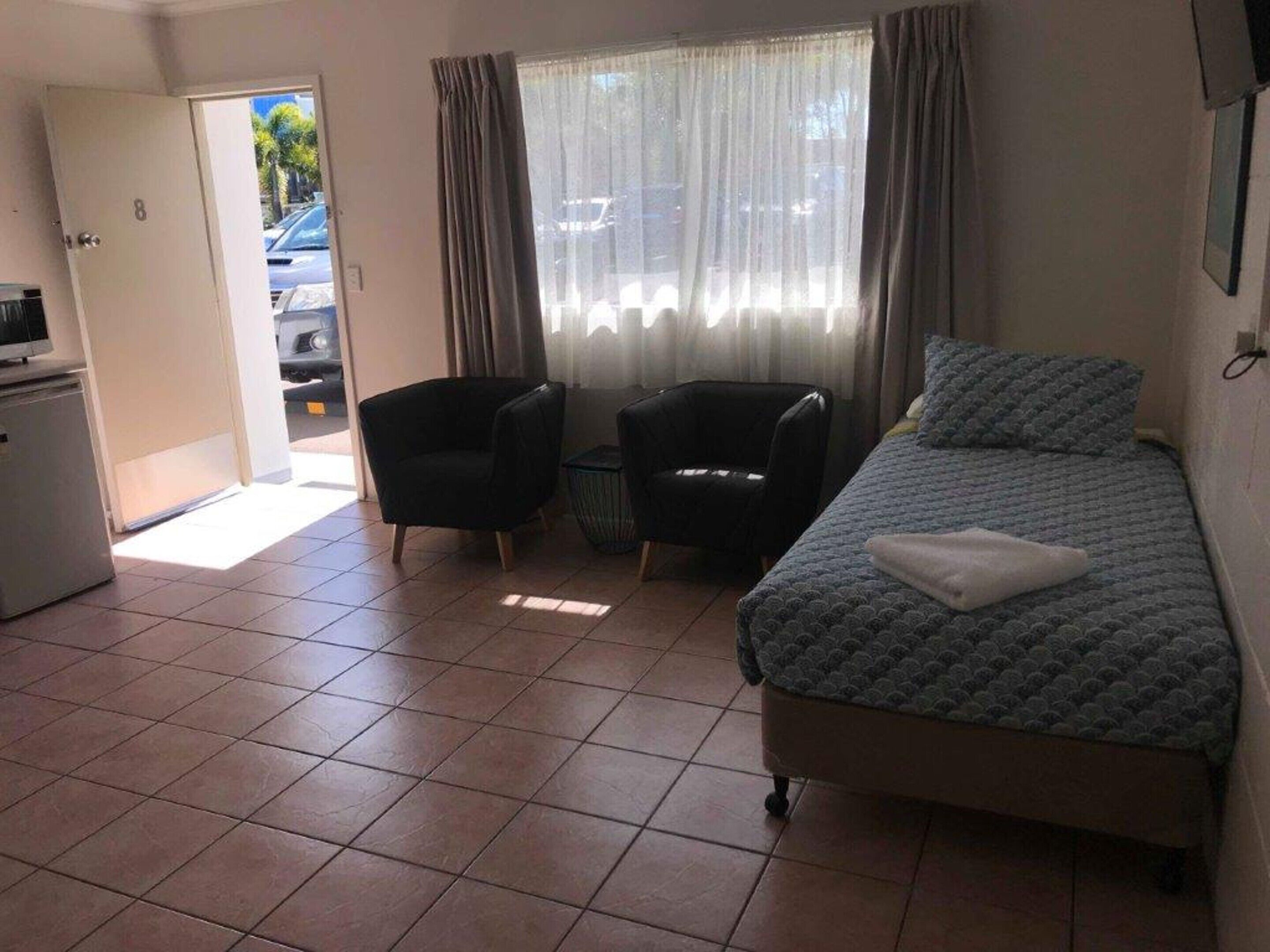 A small, clean room with a bed, two chairs, a desk, and a window with curtains.