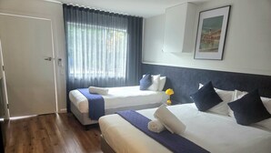 Habitación de hotel con dos camas, una puerta, una ventana con cortinas, un cuadro enmarcado en la pared y suelo de madera.
