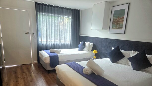 A hotel room with two beds, a door, a window with curtains, a framed picture on the wall, and a wooden floor.