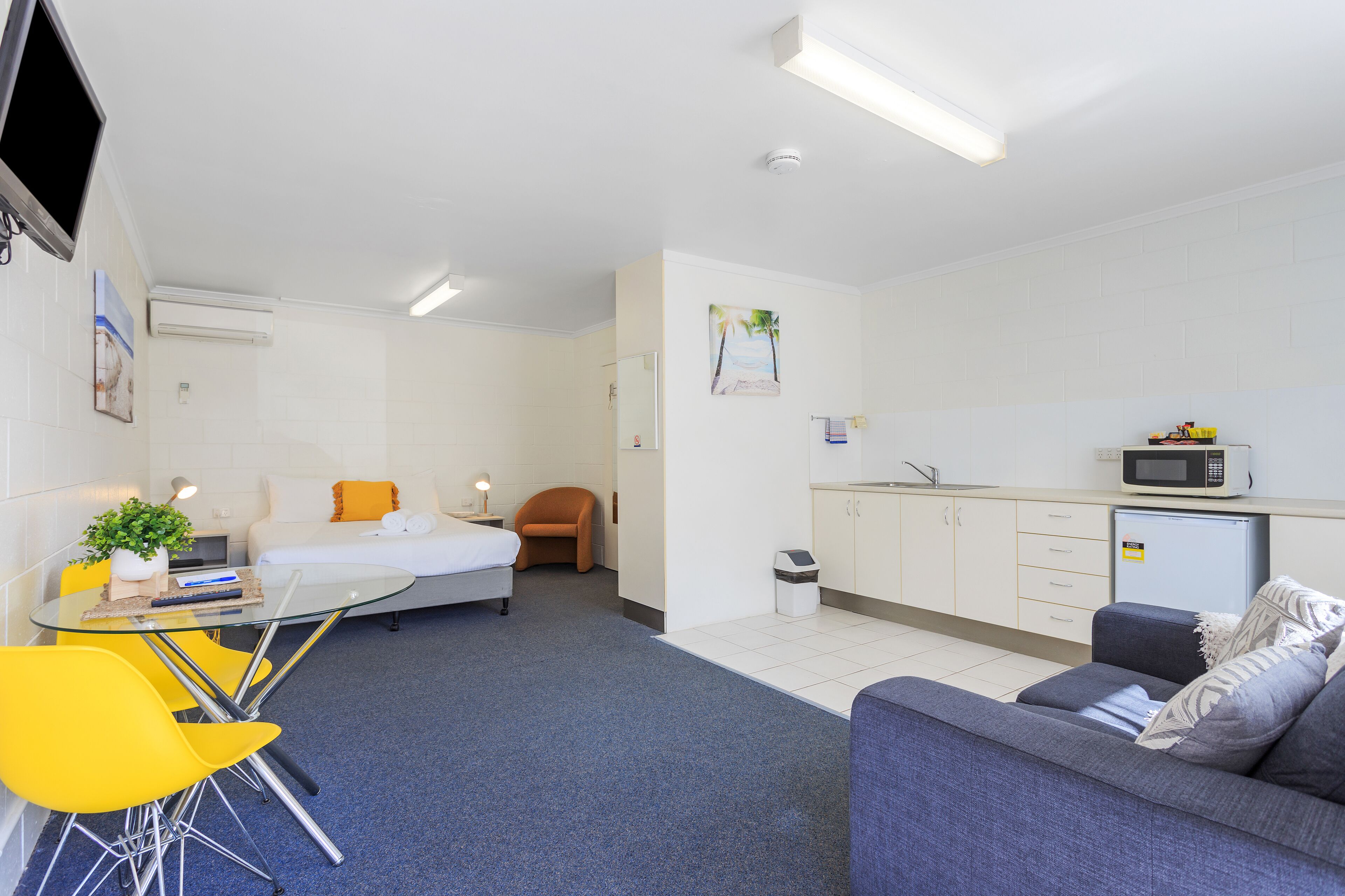 A modern, well-lit room with a bed, a sofa, a small dining table with chairs, a microwave, and a trash bin.
