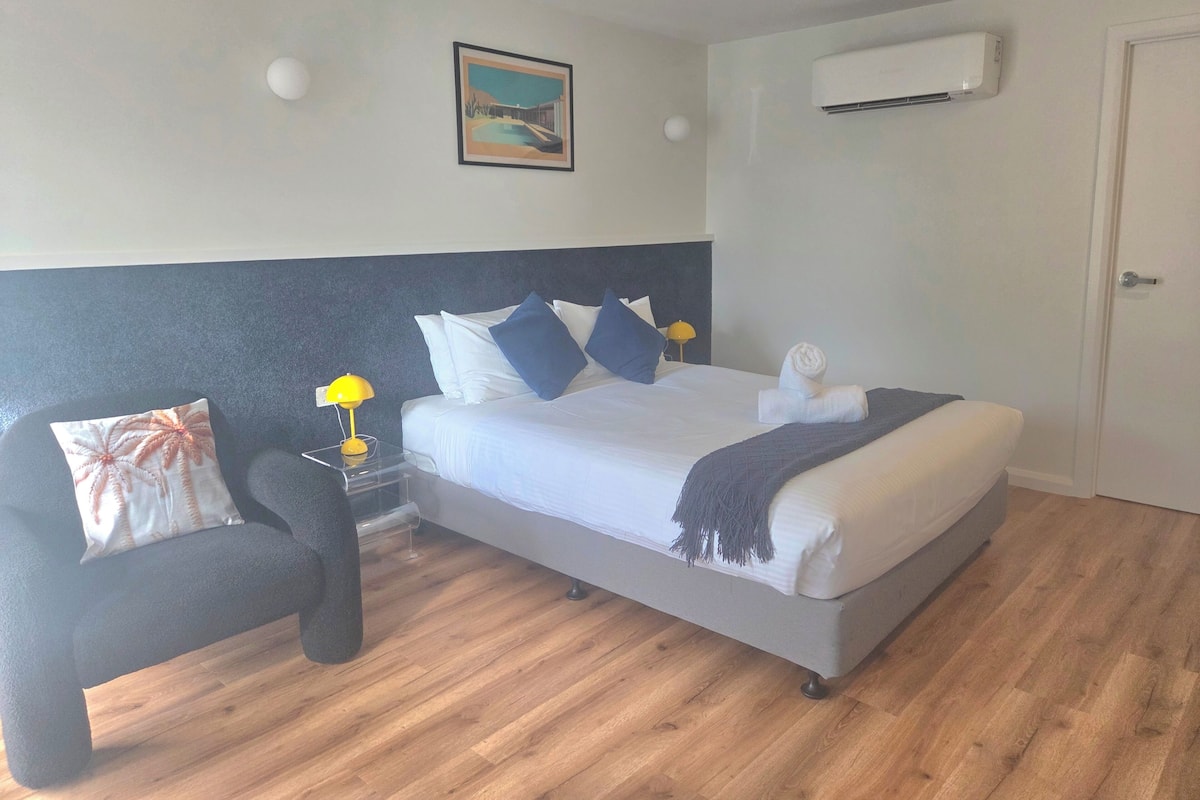 Una habitación de hotel con una cama, una mesita de noche con lámpara, aire acondicionado de pared y un cuadro.