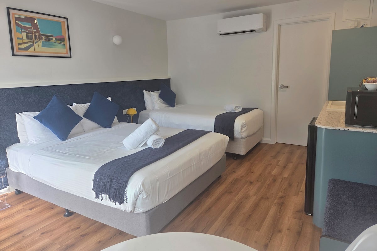 Habitación de hotel con dos camas, un cuadro en la pared, un equipo de aire acondicionado y una pequeña zona de kitchenette.
