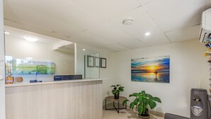 A reception area with a wooden reception desk, a computer, framed certificates, a painting of a sunset, and two potted plants.