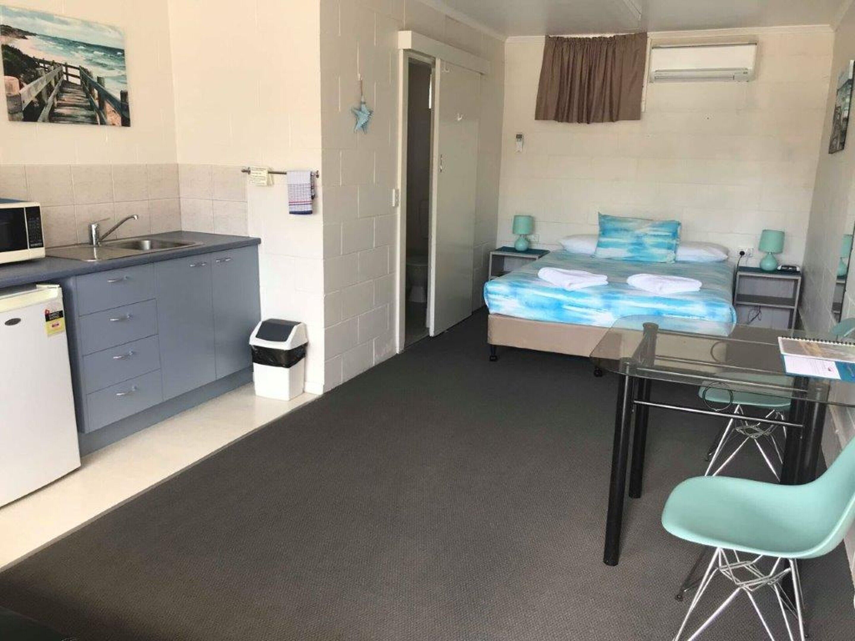 A compact hotel room with a kitchenette, a bed with a blue ocean-themed bedspread, a small table with two chairs, and a trash bin.