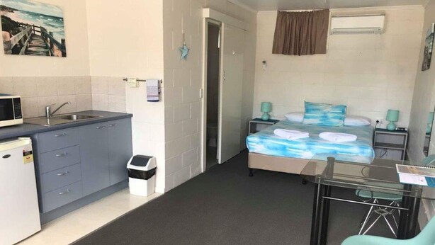 Habitación de hotel compacta con kitchenette, una cama con colcha de motivos marinos azules, una pequeña mesa con dos sillas y un cesto de basura.