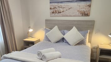 Un lit bien fait, recouvert d’une couverture blanche et agrémenté d’oreillers, avec un tableau représentant une scène de plage au-dessus, et deux lampes de chevet de chaque côté.