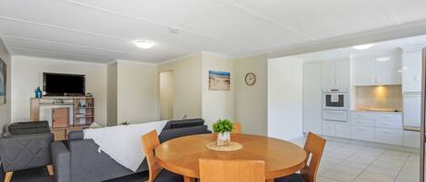 A modern living room with a dining table, grey sofas, and a kitchen area with white cabinets.