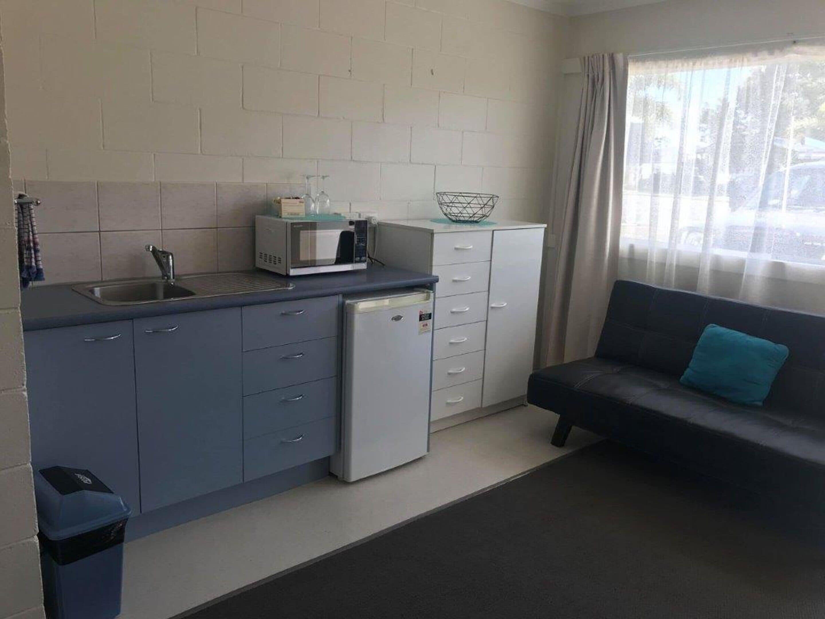A compact kitchen with a sink, microwave, and small refrigerator. There is a dining area with a sofa and a window with curtains.