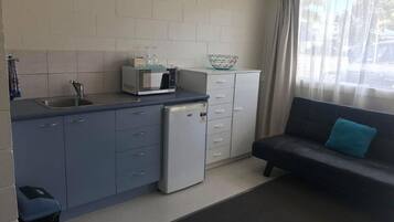 A compact kitchen with a sink, microwave, and small refrigerator. There is a dining area with a sofa and a window with curtains.