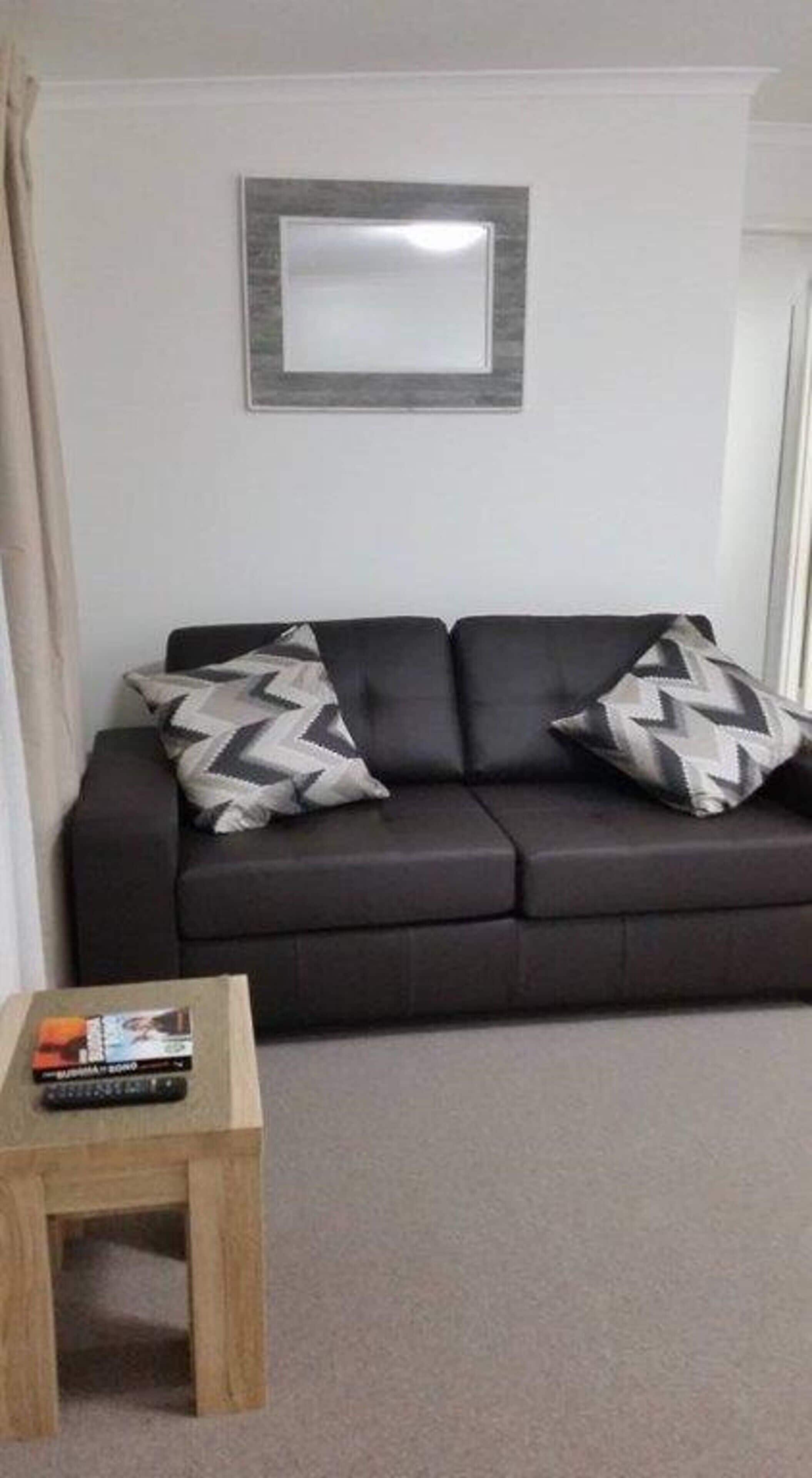 Een moderne woonkamer met een donkere bank, twee kussens met een patroon, een klein houten bijzettafeltje met boeken, en een lijst met een foto aan de muur.