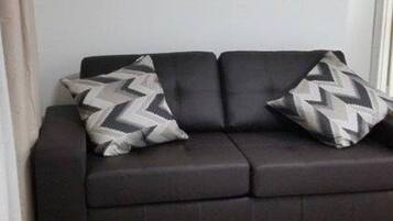 Een moderne woonkamer met een donkere bank, twee kussens met een patroon, een klein houten bijzettafeltje met boeken, en een lijst met een foto aan de muur.