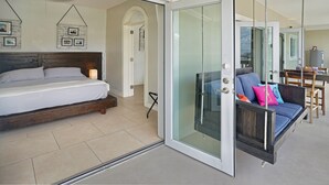 A bedroom with a bed, a glass door leading to a balcony, and a dining area visible through the door. - Two Sandals – A Boutique Hotel (St. Thomas)