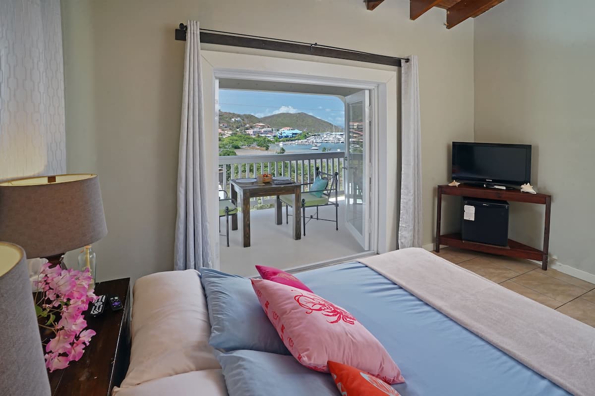 Ein Hotelzimmer mit einem Bett, einem Fernseher auf einem Holztischchen, einem Balkon mit Tisch und Stühlen sowie Blick auf einen Jachthafen und Gebäude.
