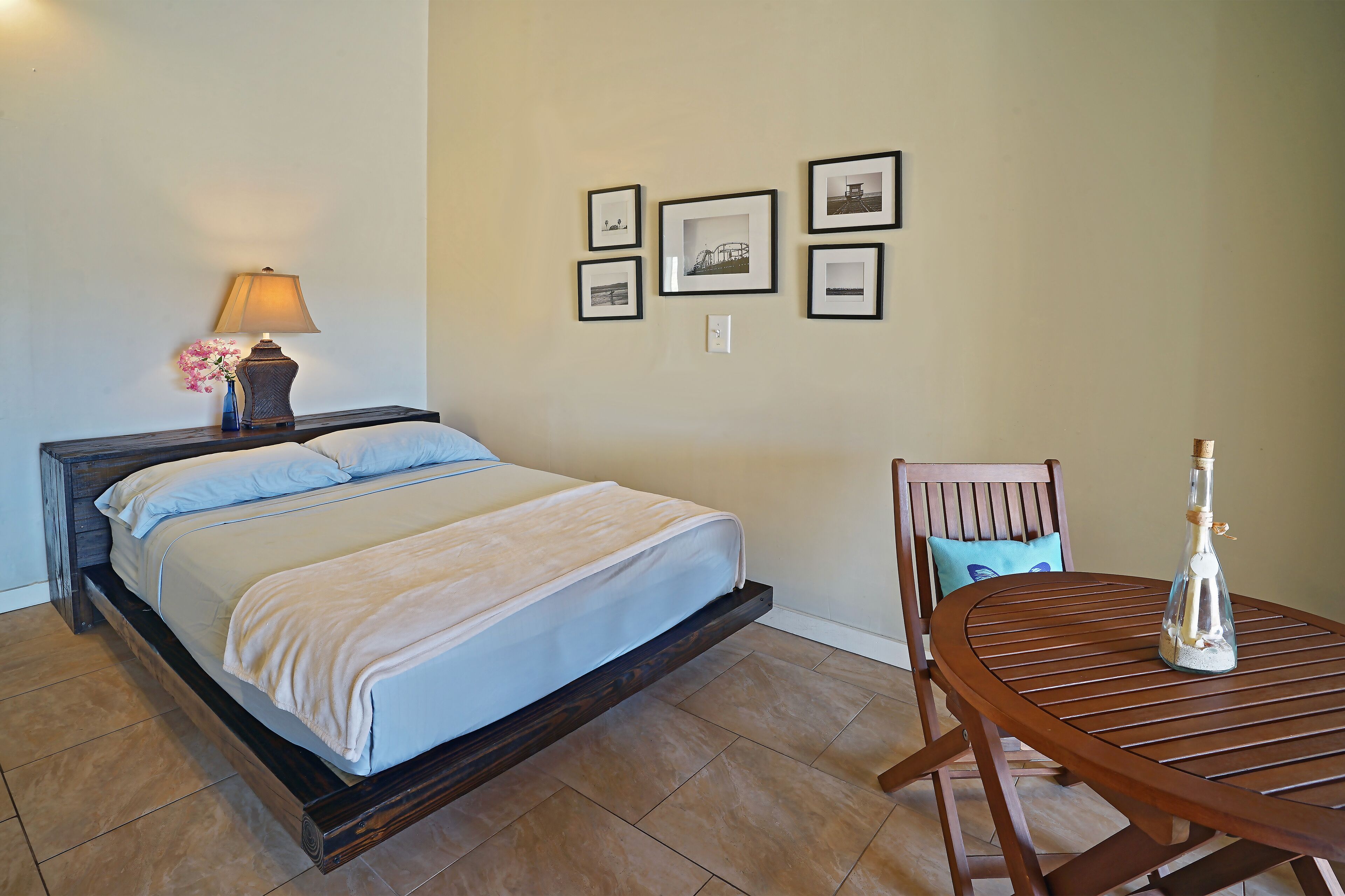 Un dormitorio con cama, mesita de noche con lámpara, una silla y una mesa con una botella.