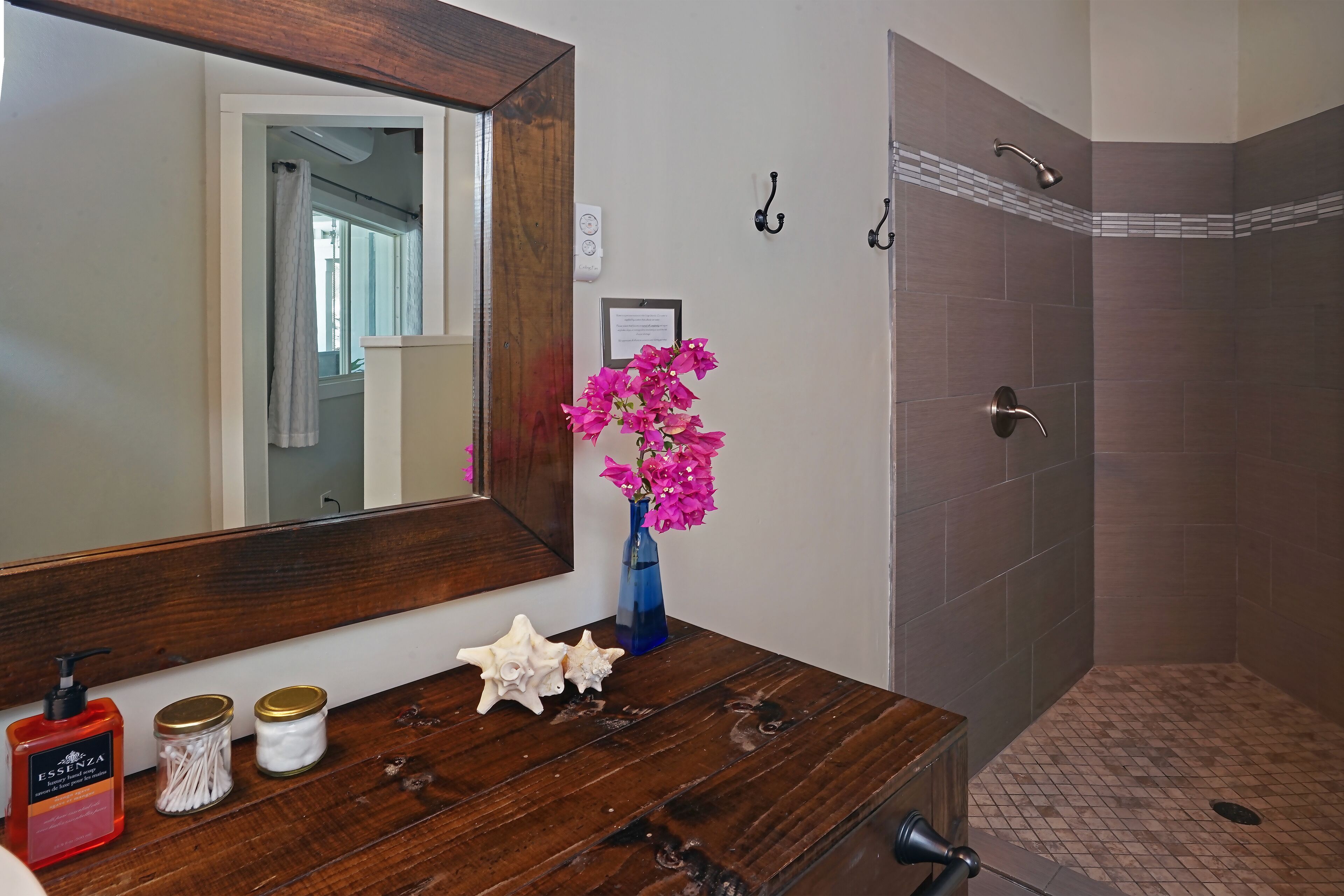 A bathroom with a wooden vanity, a large mirror, a shower with a tiled floor, and a vase with pink flowers.