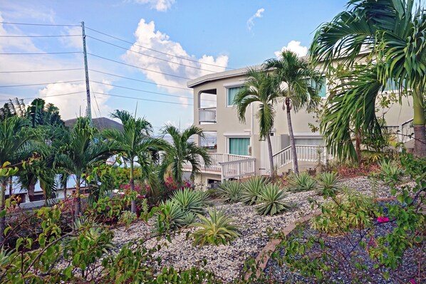 Un edificio moderno con balcón, rodeado de plantas tropicales y un jardín rocoso.