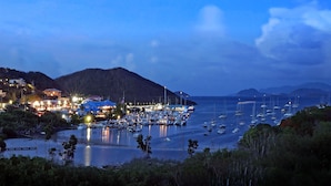 A marina at dusk with boats docked, a waterfront town with buildings, and hills in the background. - Two Sandals – A Boutique Hotel (St. Thomas)