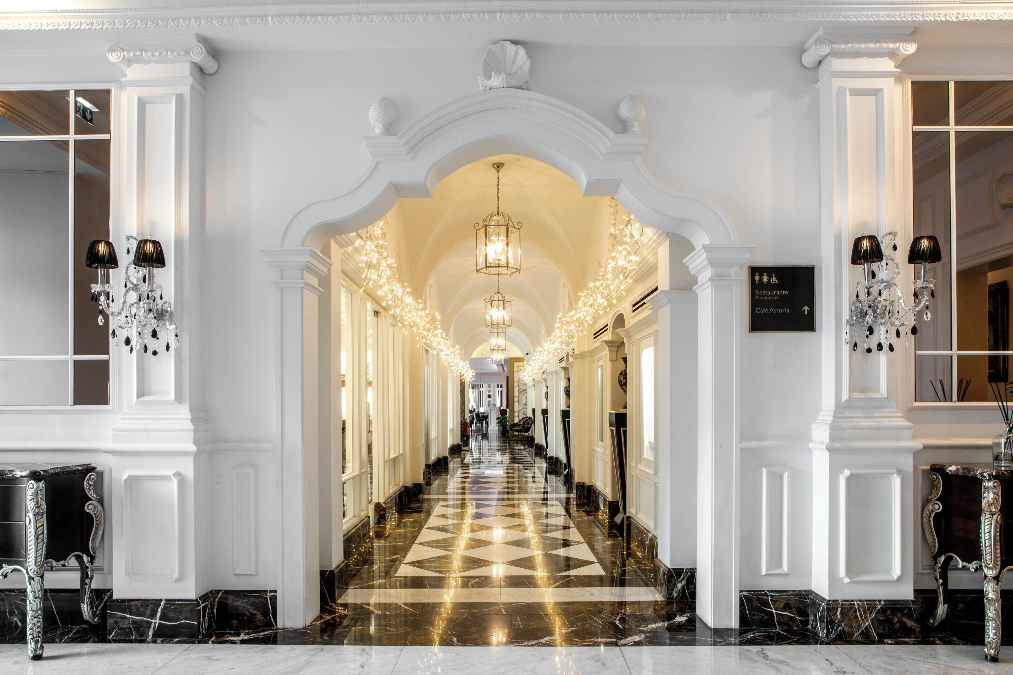 Un amplio pasillo con suelo de mármol a cuadros, columnas blancas y candelabros.