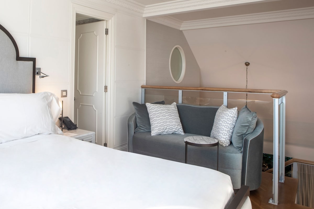 A bedroom with a bed, a bedside table, a telephone, and a grey sofa with patterned cushions.