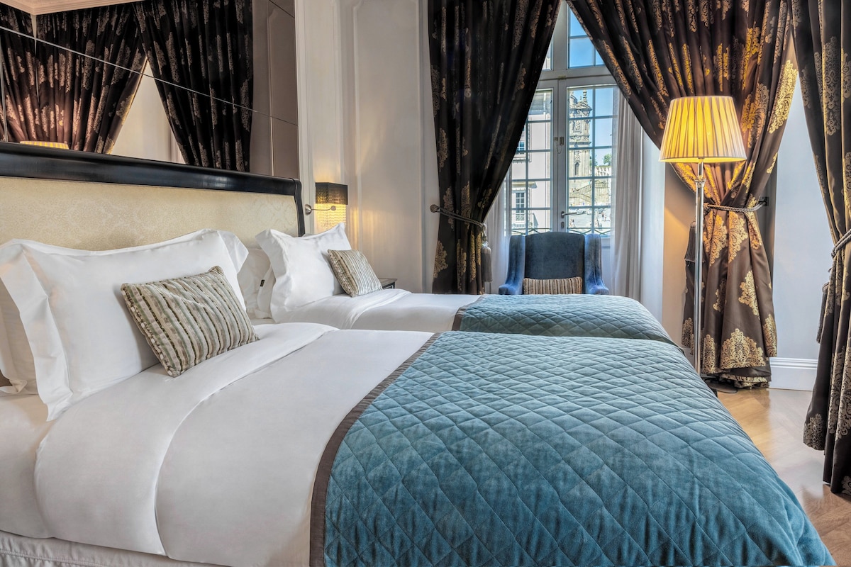 A hotel room with two beds, dark curtains, a lamp, and a view of a building through the window.
