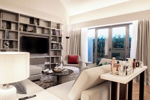 A modern living room with a flat-screen TV, comfortable seating, a coffee table, and a view of greenery through large windows.