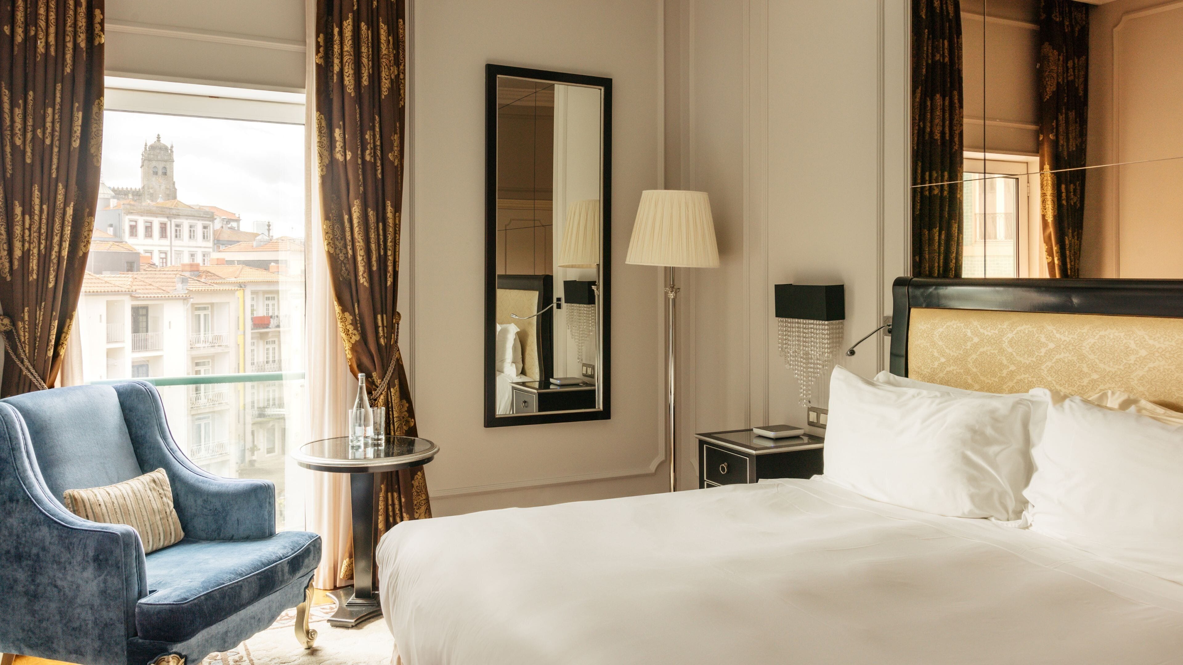 A hotel room with a bed, a chair, a nightstand, a lamp, and a view of a cityscape.