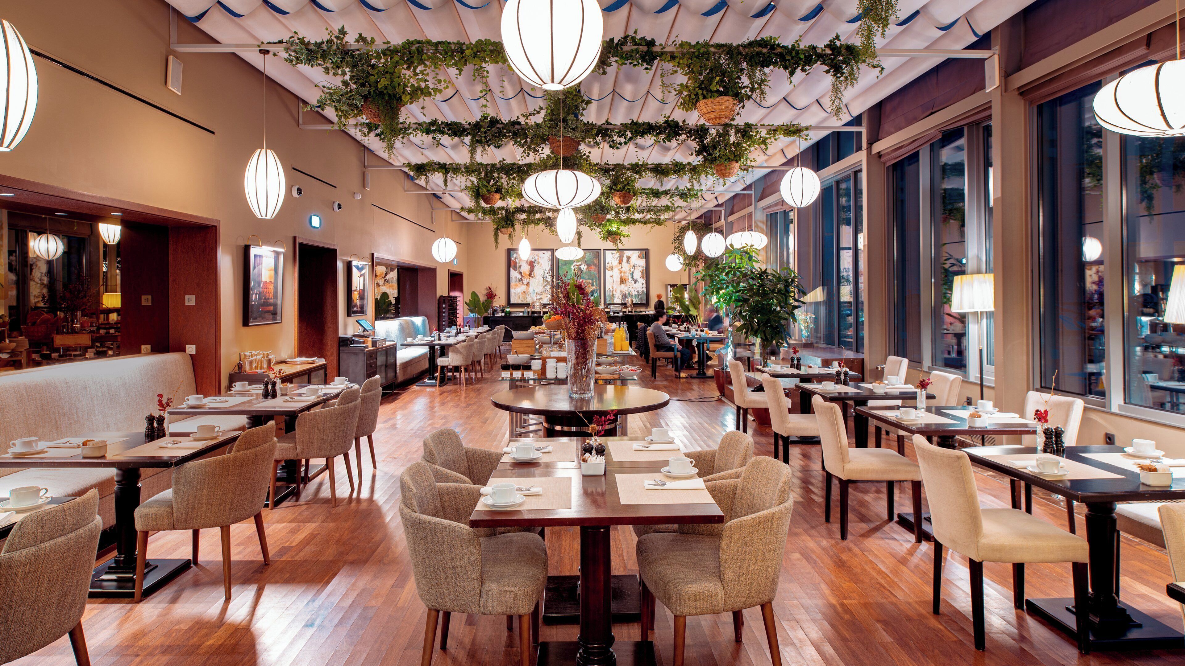 Un restaurante amplio con varias mesas y sillas, ventanas grandes y plantas colgantes.