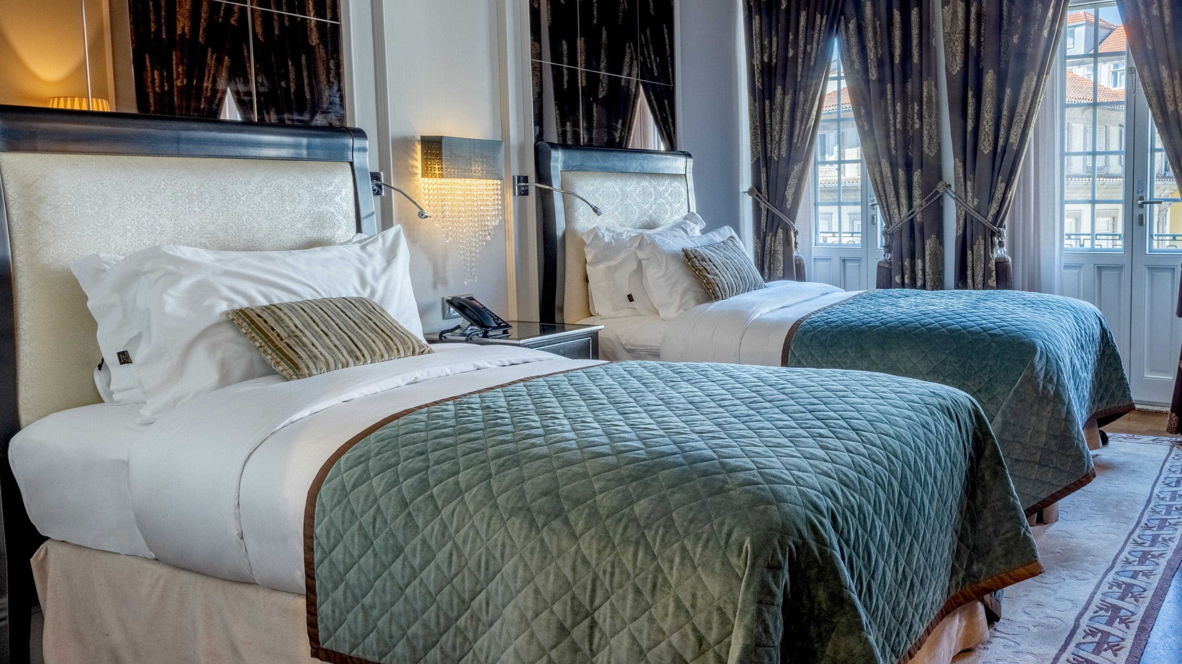 A hotel room with two beds, each with a headboard, a bedside table with a lamp, and a view of the outside through large windows.