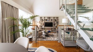 A modern living room with a flat-screen TV, a glass coffee table, and a staircase with glass railings.