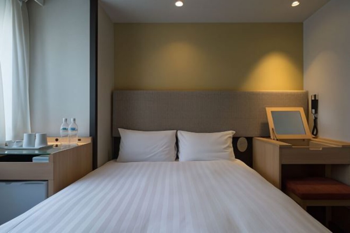 A hotel room with a bed, a bedside table with a lamp, a mirror, and a small desk with a computer.