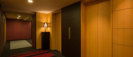 A hotel hallway with wooden doors, a checkered wall pattern, a lamp, and a red and black striped carpet.
