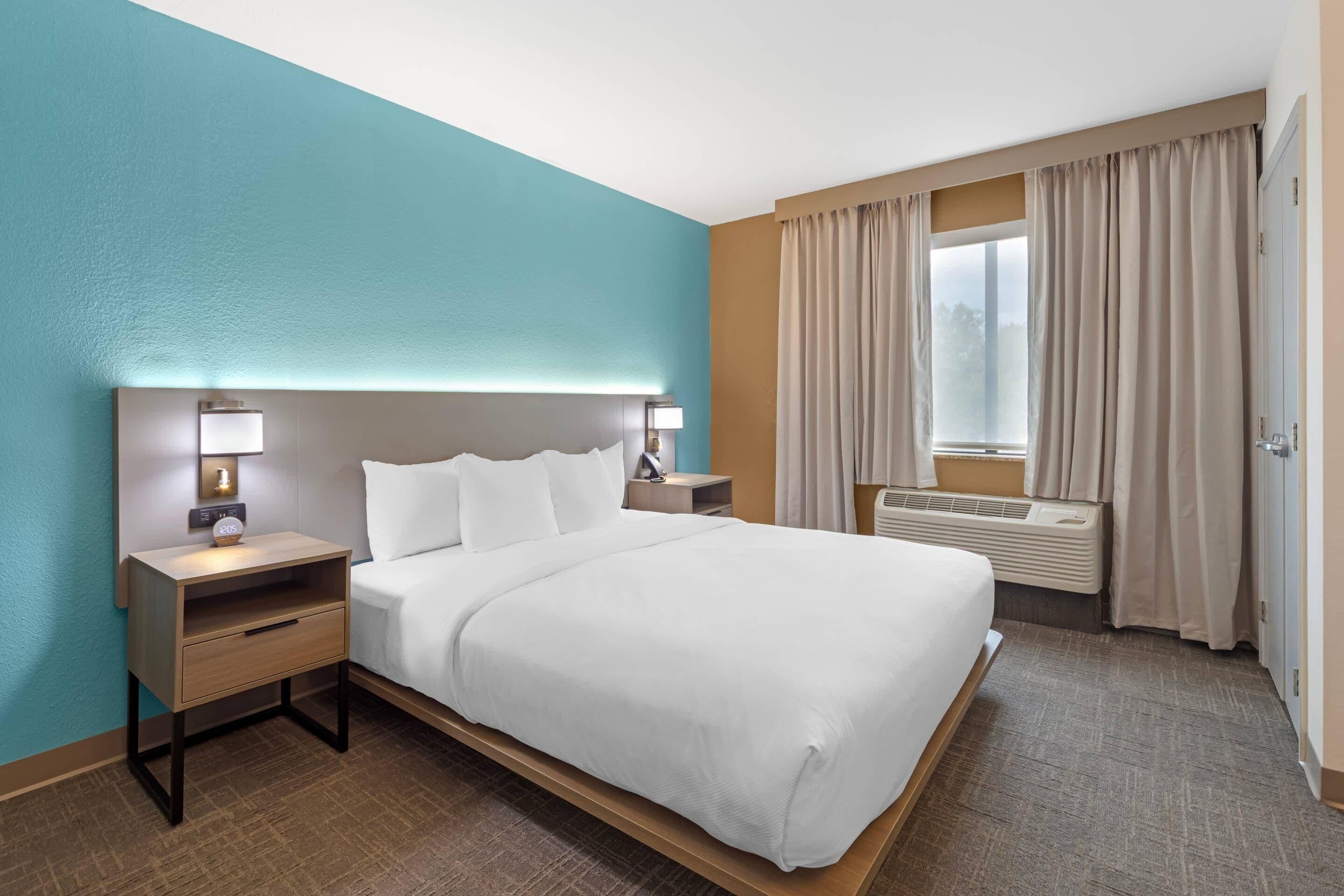 A hotel room with a bed, a bedside table with a lamp, a nightstand, a window with curtains, and an air conditioning unit.