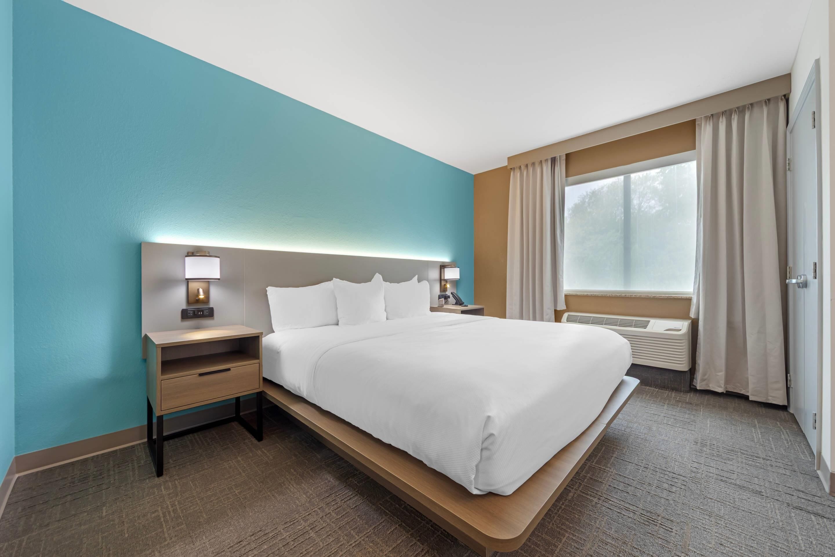 A hotel room with a large bed, a bedside table with a lamp, a window with curtains, and a wall painted in teal.