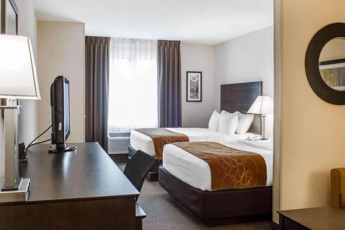 A hotel room with a desk, two beds, a TV, a mirror, and a window with curtains.