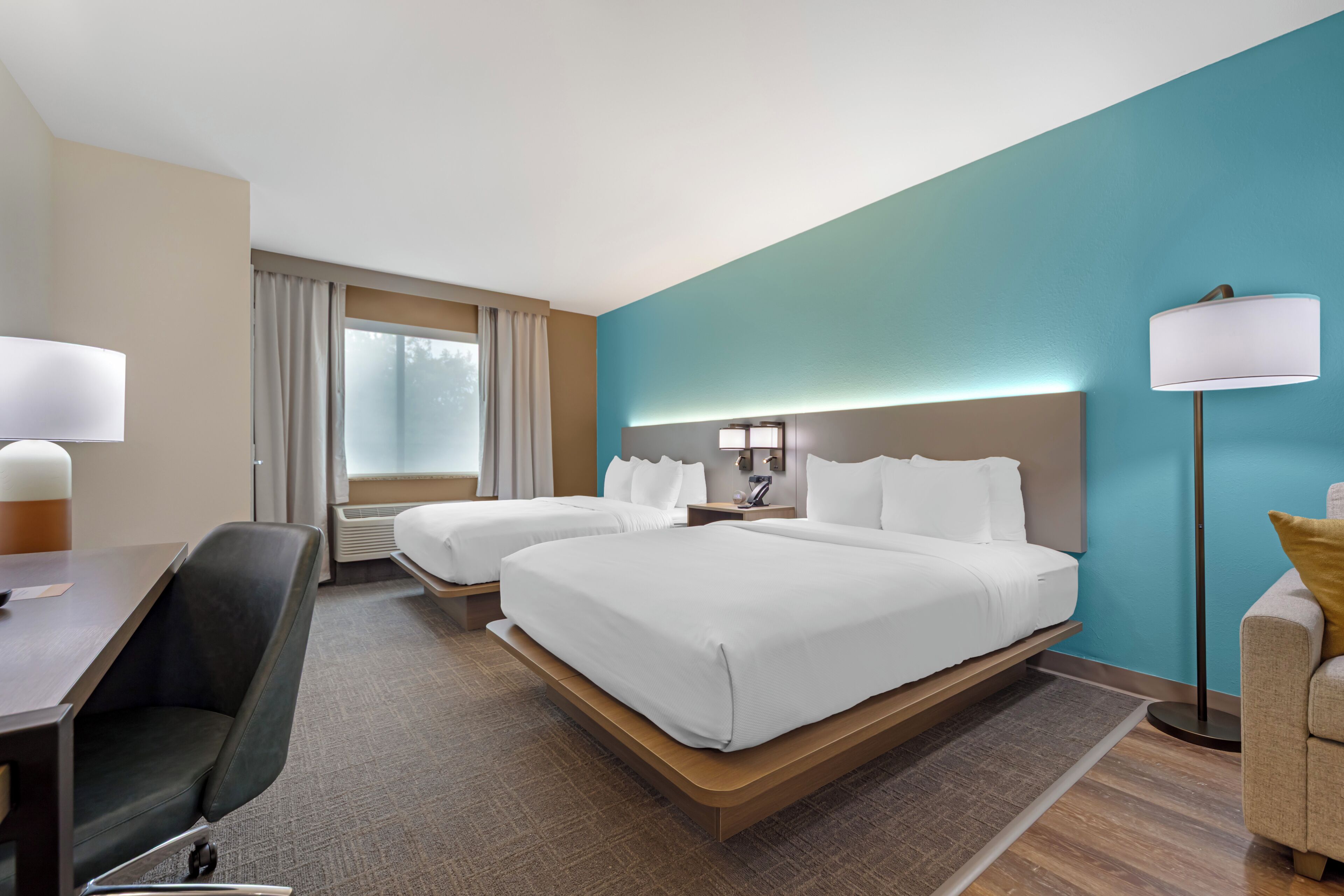A hotel room with two beds, a desk, a chair, a lamp, and a view of the outdoors.
