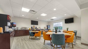 A modern dining area with wooden tables and orange chairs, a coffee machine, and a TV mounted on the wall. - Comfort Suites Lewisburg near University (Lewisburg)