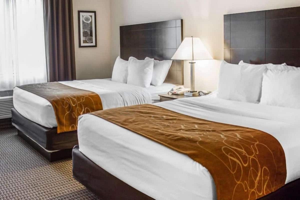 A hotel room with two beds, a bedside table with a lamp, a framed picture on the wall, and a window with curtains.