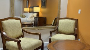 Habitación de hotel con dos sillas de madera, una mesa redonda, una cama, una mesita de noche, una lámpara y un espejo grande.