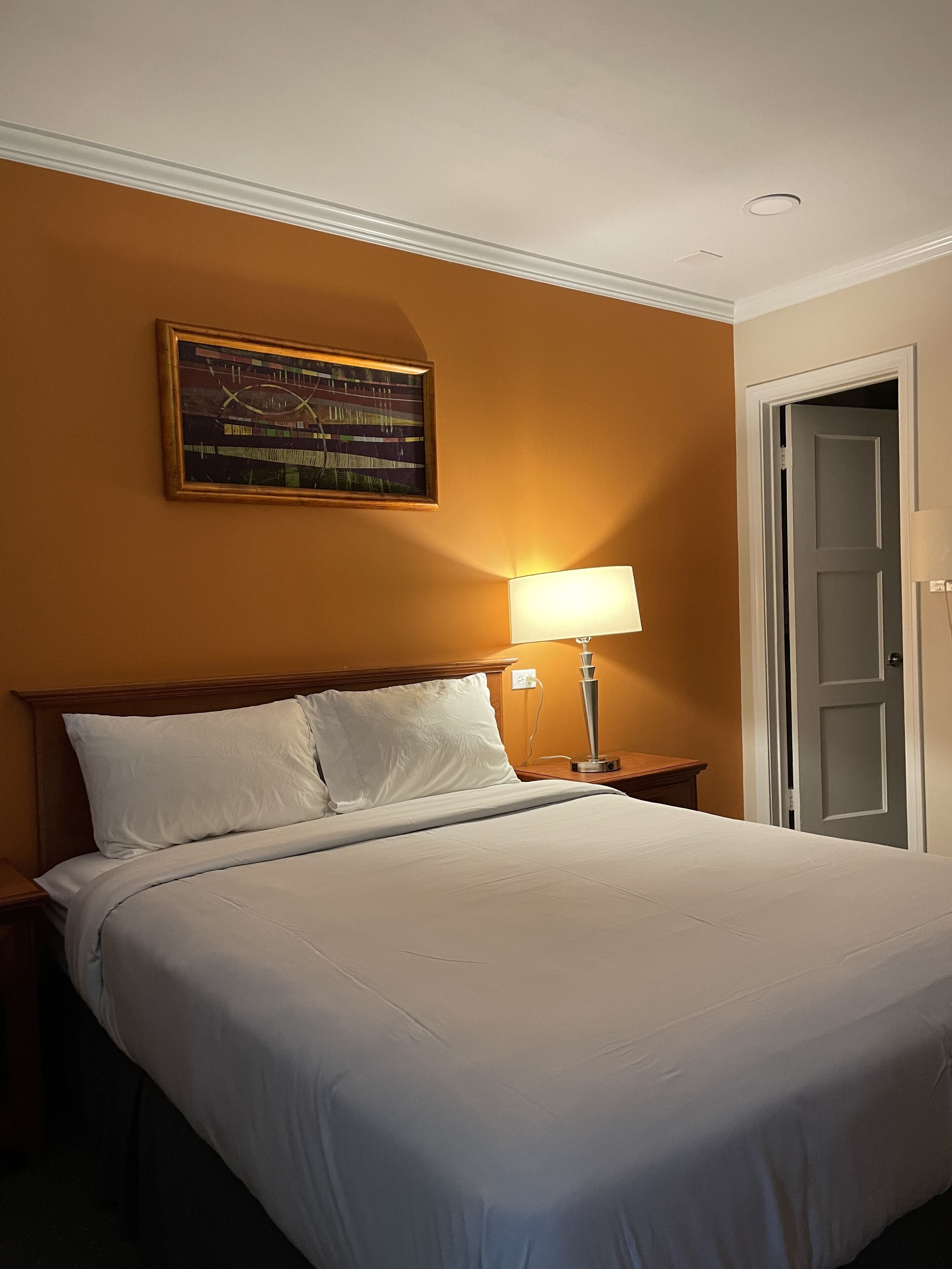 Una habitación de hotel con una cama grande, una mesita de noche con lámpara, un cuadro en la pared y una puerta.