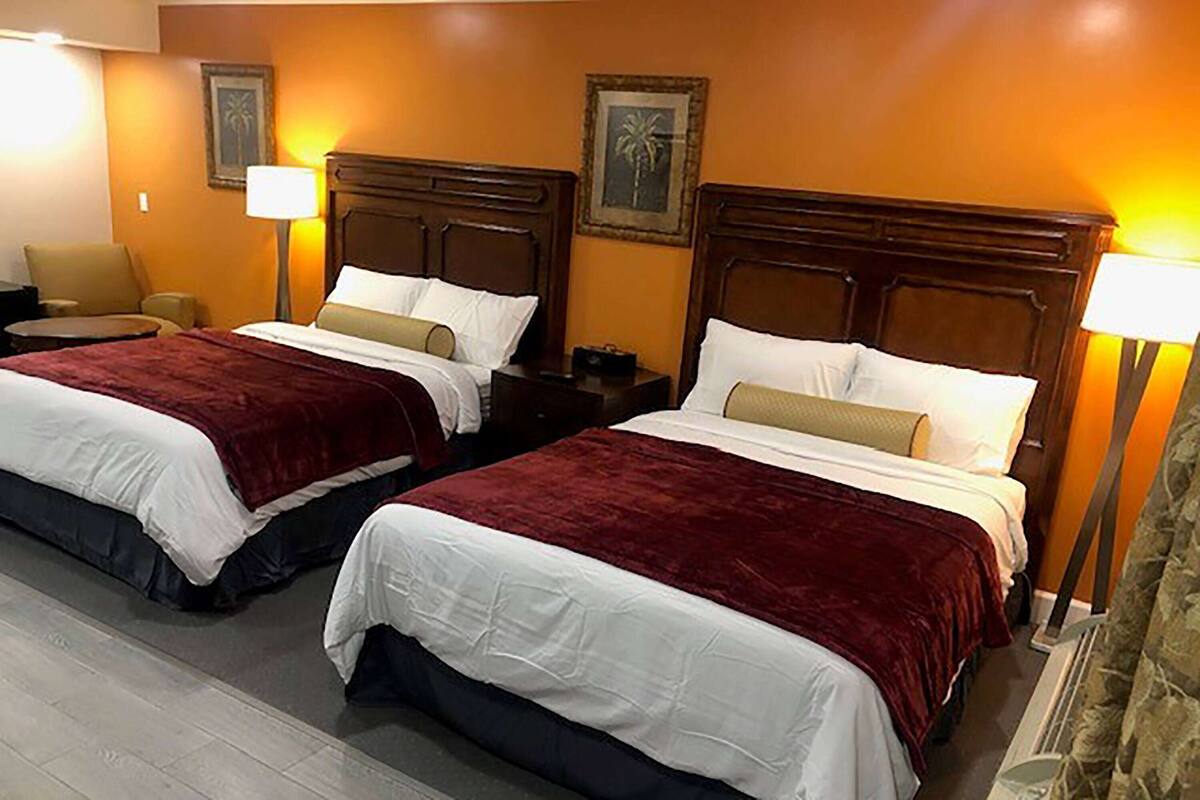 A hotel room with two beds, each with a wooden headboard, white bedding, and a red throw blanket. There are two bedside tables with lamps, a chair, and framed pictures on the wall.
