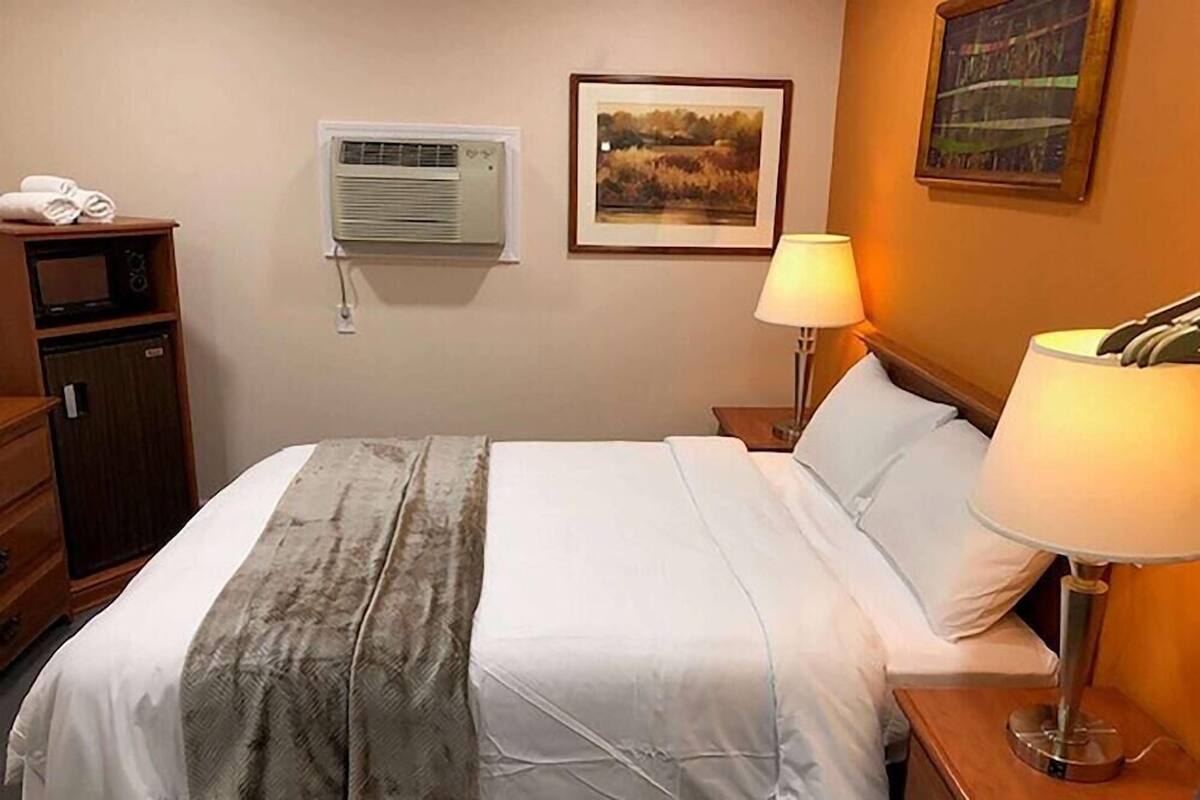 A hotel room with a bed, a bedside lamp, an air conditioning unit, a framed picture on the wall, and a wooden dresser.