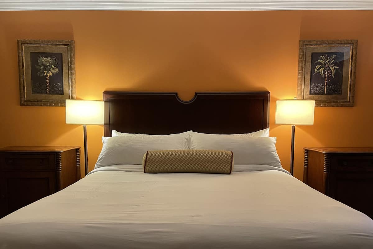 A large bed with white linens and a wooden headboard, flanked by two nightstands with lamps, in a room with orange walls and framed artwork.