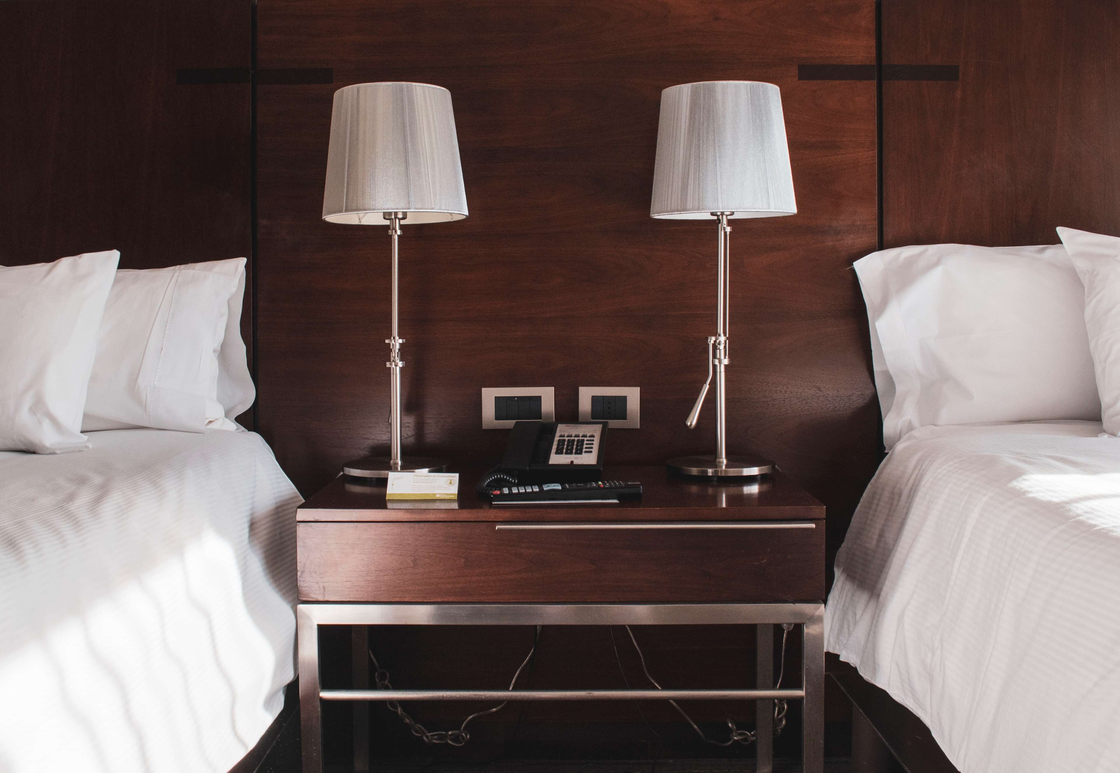 Habitación de hotel con dos camas, una mesita de noche de madera con un teléfono y un control remoto, y dos lámparas fijadas a la pared.