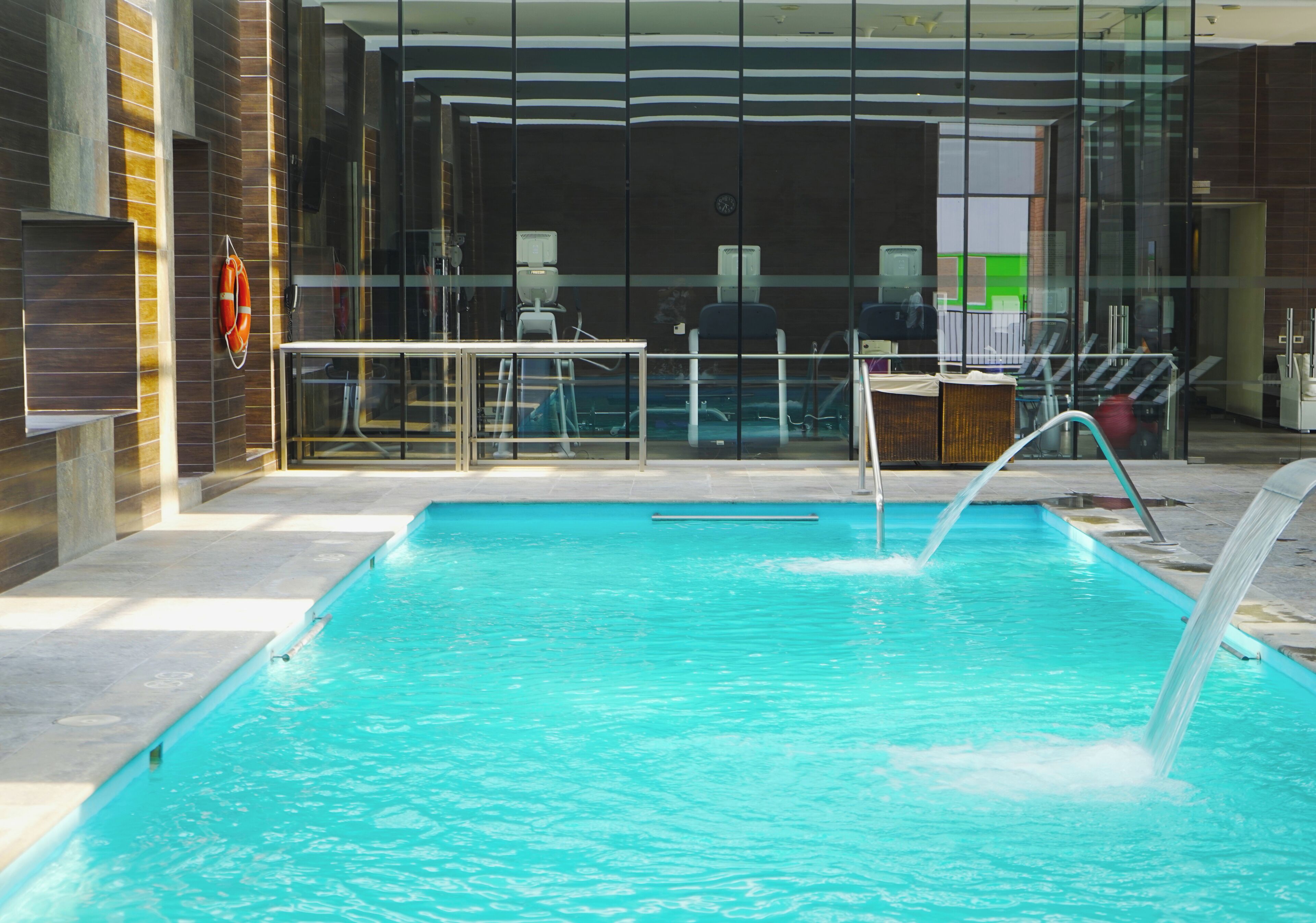 A swimming pool with a water fountain, surrounded by a glass enclosure, with a view of a modern building in the background.