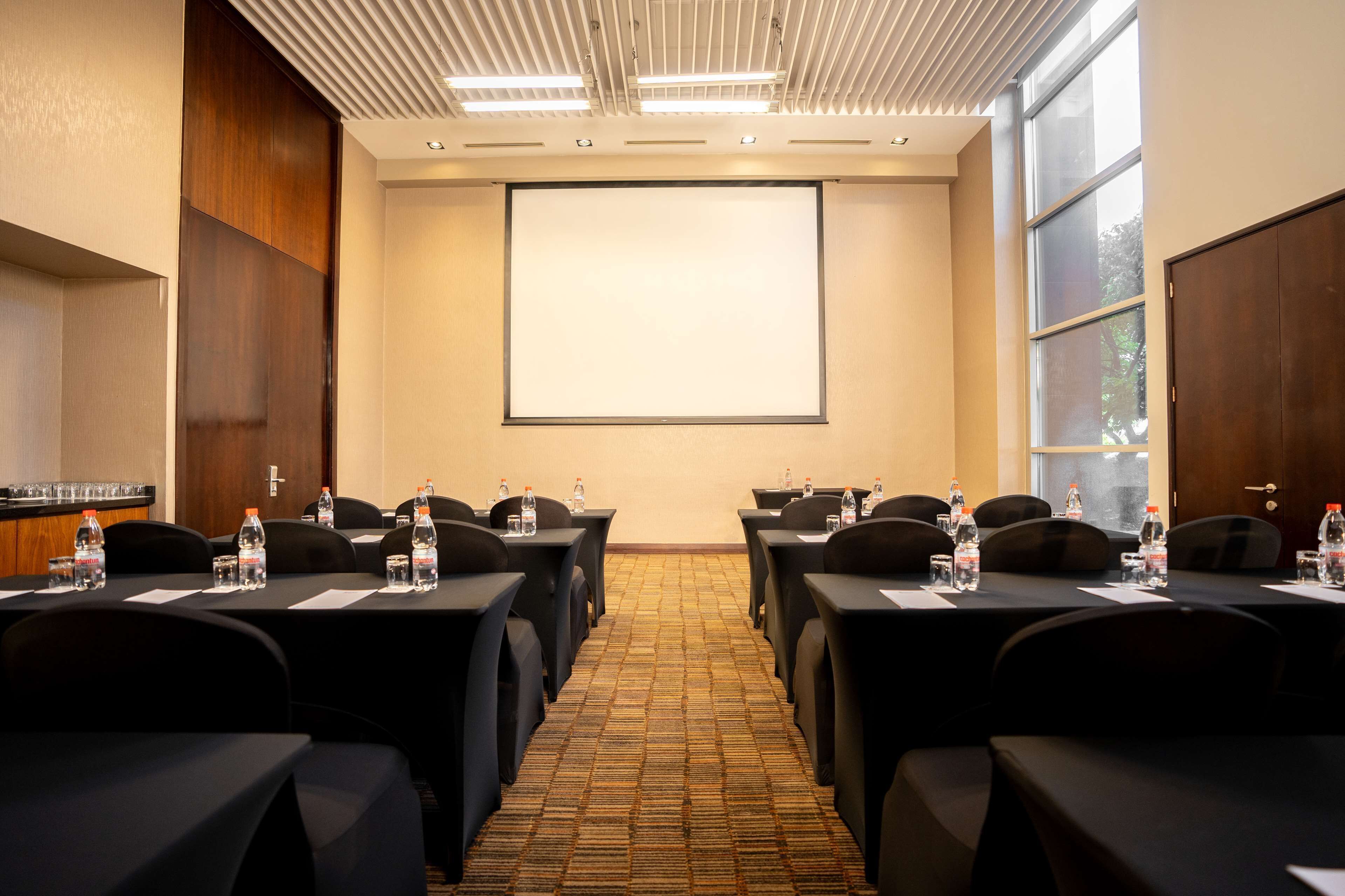 Un salón de conferencias con una gran pantalla de proyección, mesas negras con botellas de agua y piso alfombrado.