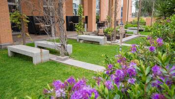 An outdoor patio area with seating arrangements, a brick building in the background, and a grassy lawn with purple flowers in the foreground.