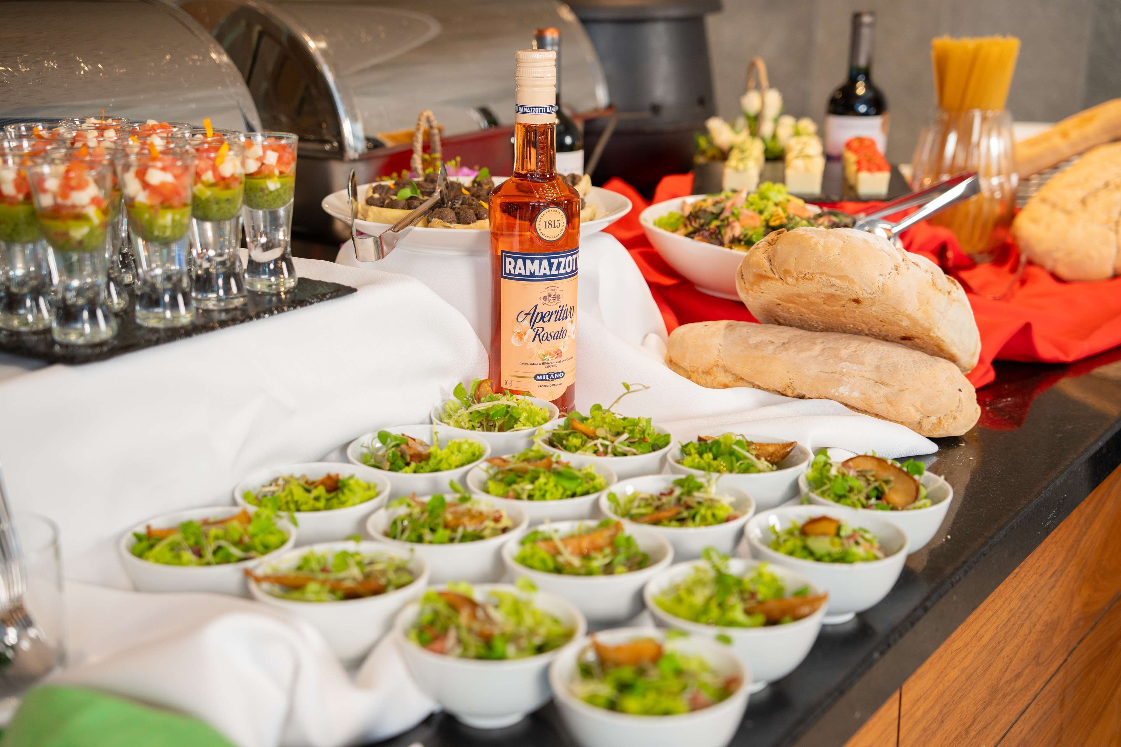 Una mesa de buffet con diversos platos, incluyendo ensaladas y pan, y una botella de Ramazzotti Aperitivo Rosato.