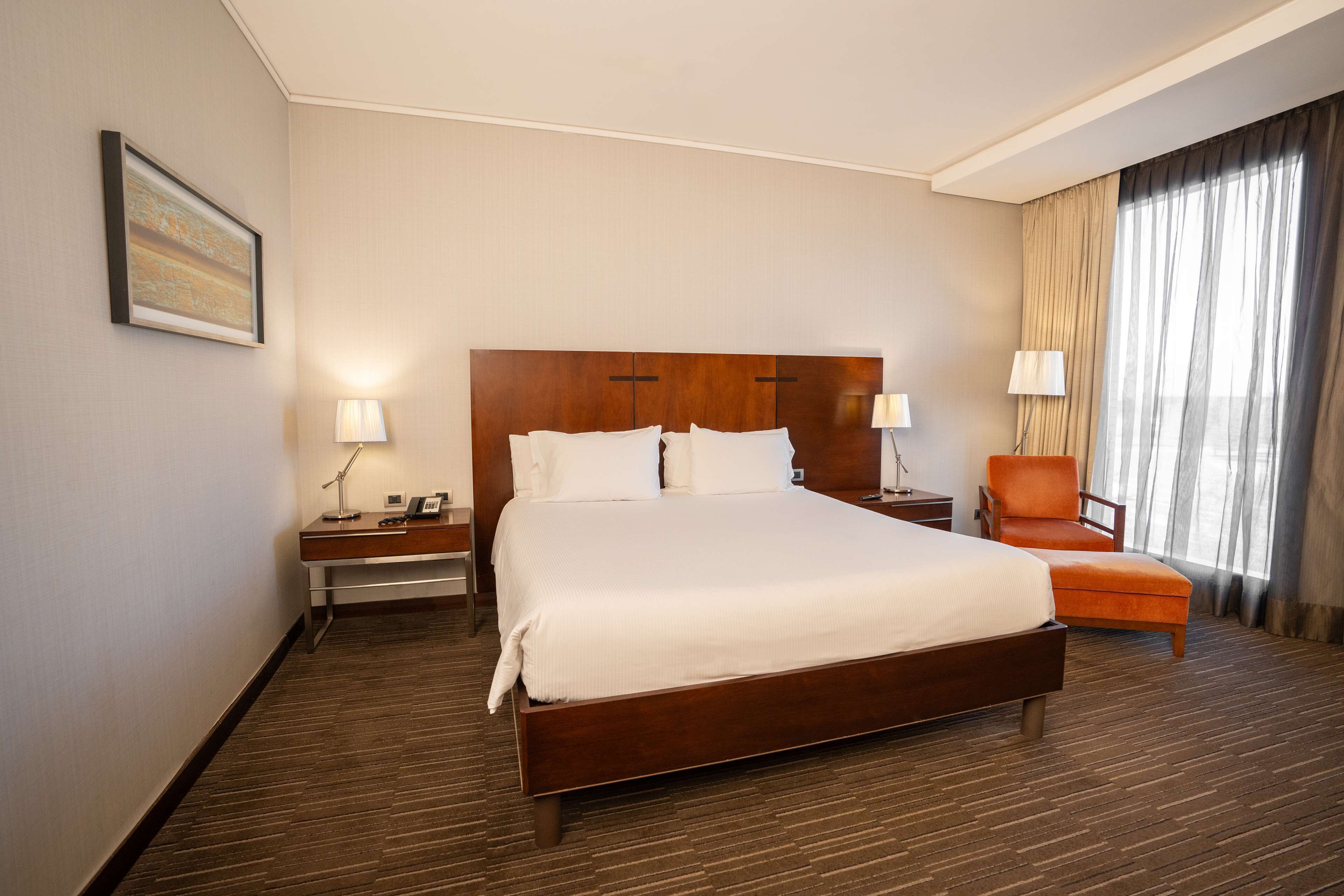 A hotel room with a large bed, two bedside tables with lamps, a chair, and a window with curtains.