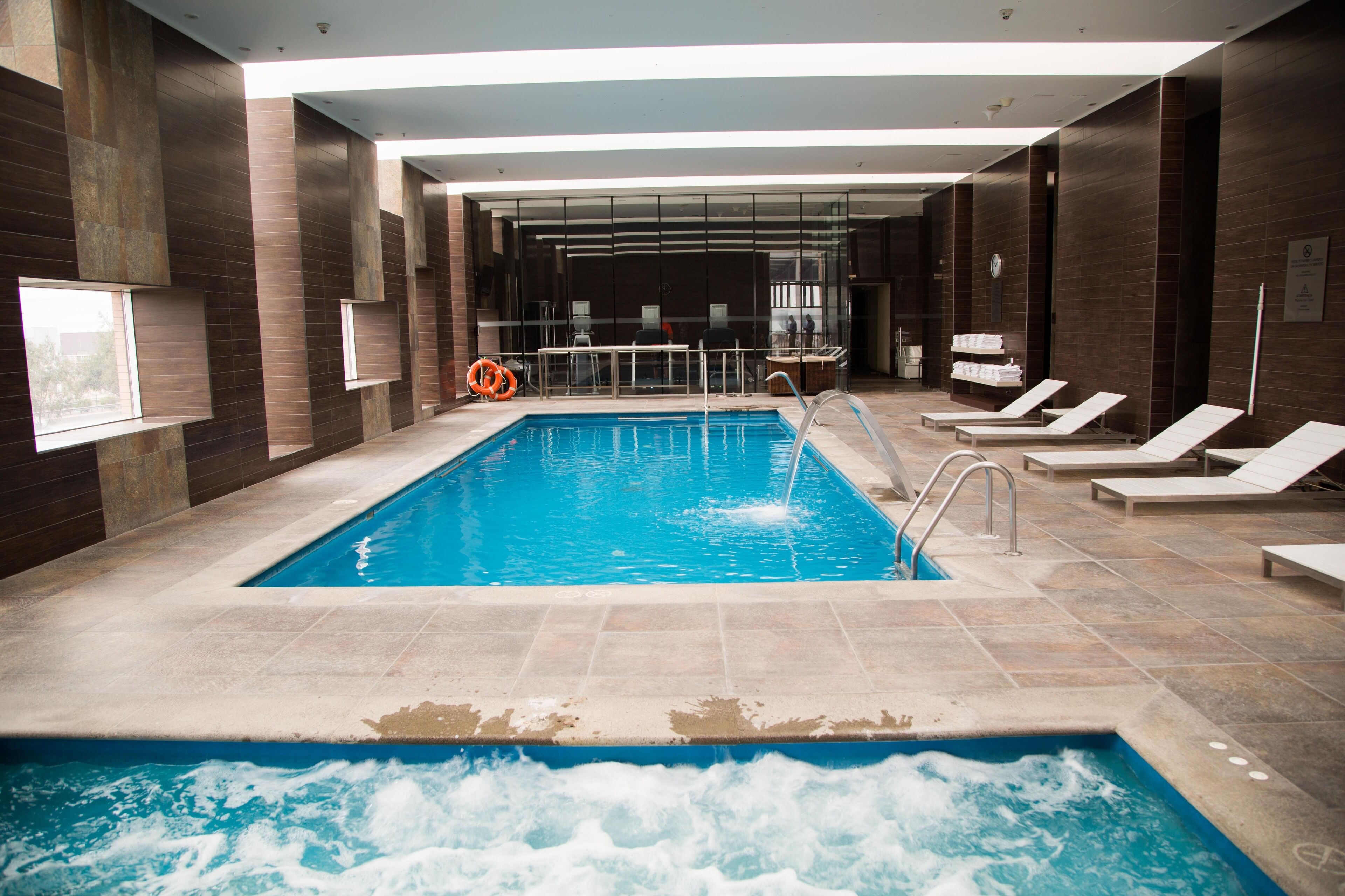 A swimming pool with a separate hot tub area, lounge chairs, and a glass-enclosed spa room.