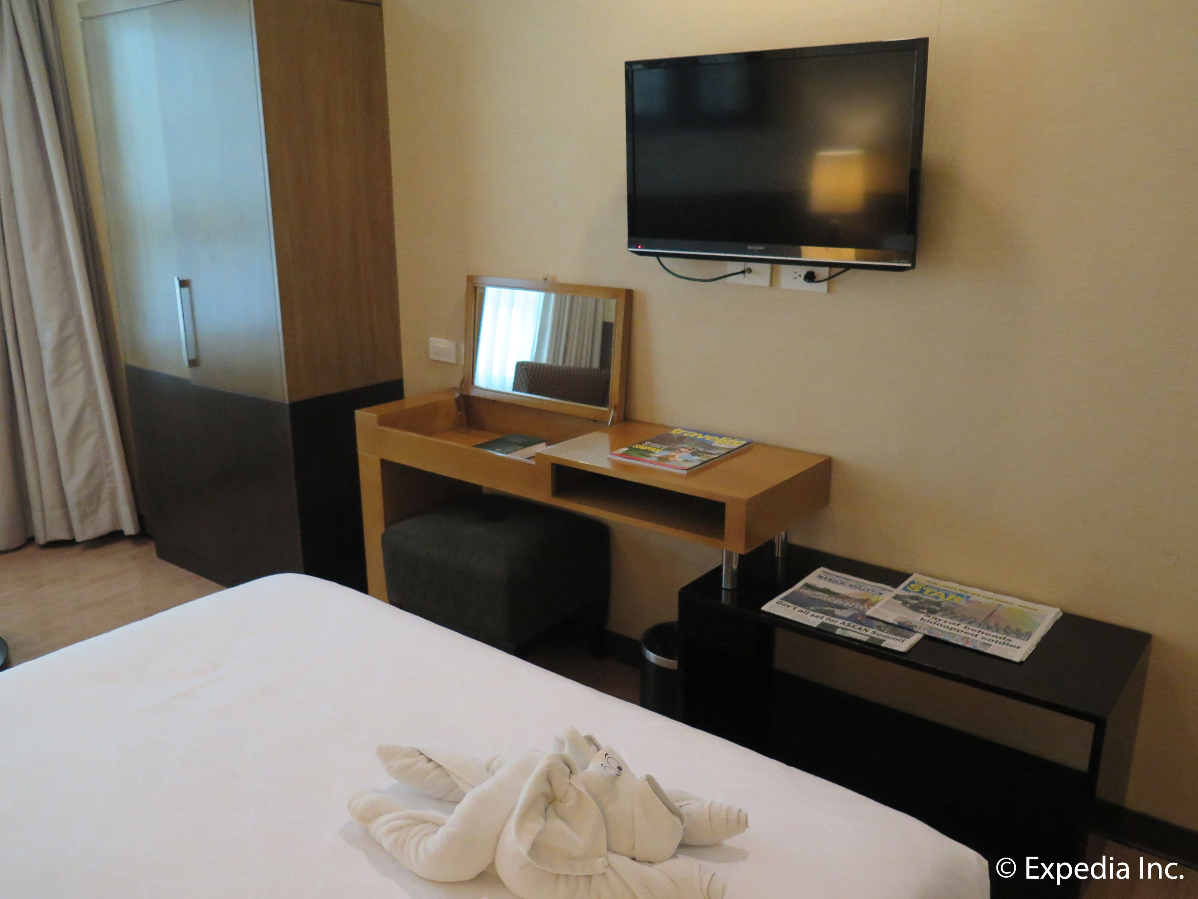 Habitación de hotel con cama, escritorio con silla, televisión montada en la pared y ventana con cortinas.